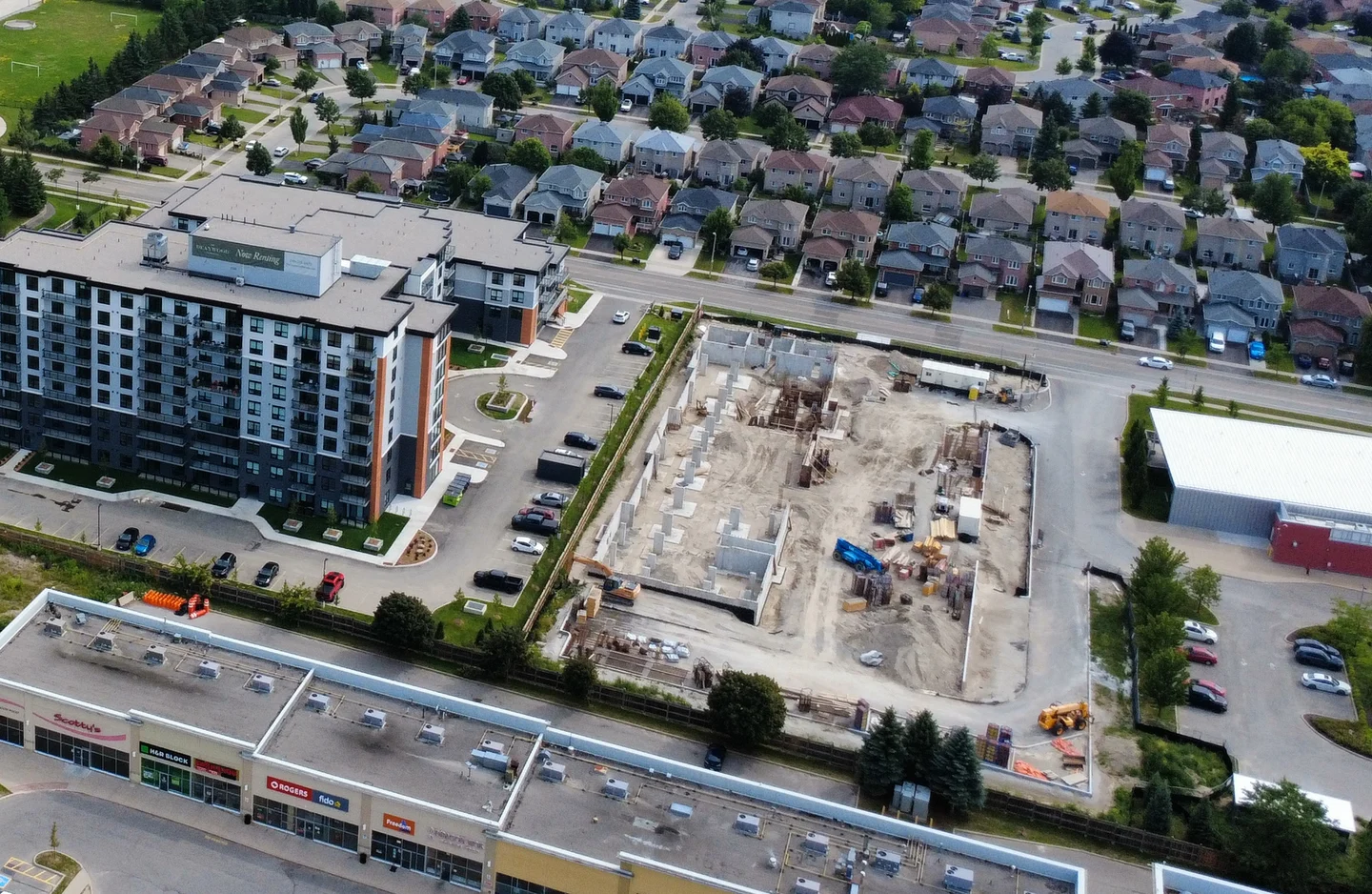 Aerial view of a completed rental apartment building, a second rental building under construction and a commercial plaza separated by a continuous fence.